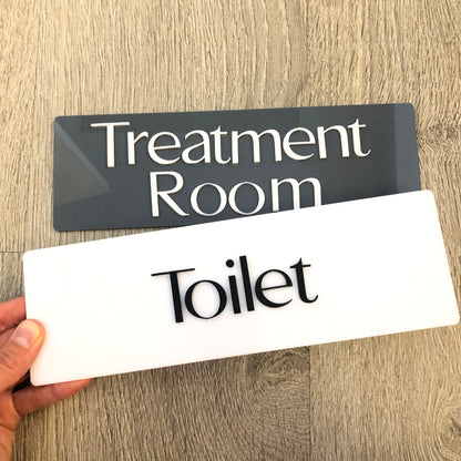 Two signs, one black with 'Treatment Room' and one white with 'Toilet', held by a hand on a wooden surface.
