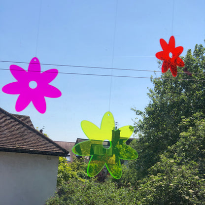 Colorful flower-shaped objects hanging on the window against a blue sky with trees and buildings in the background.