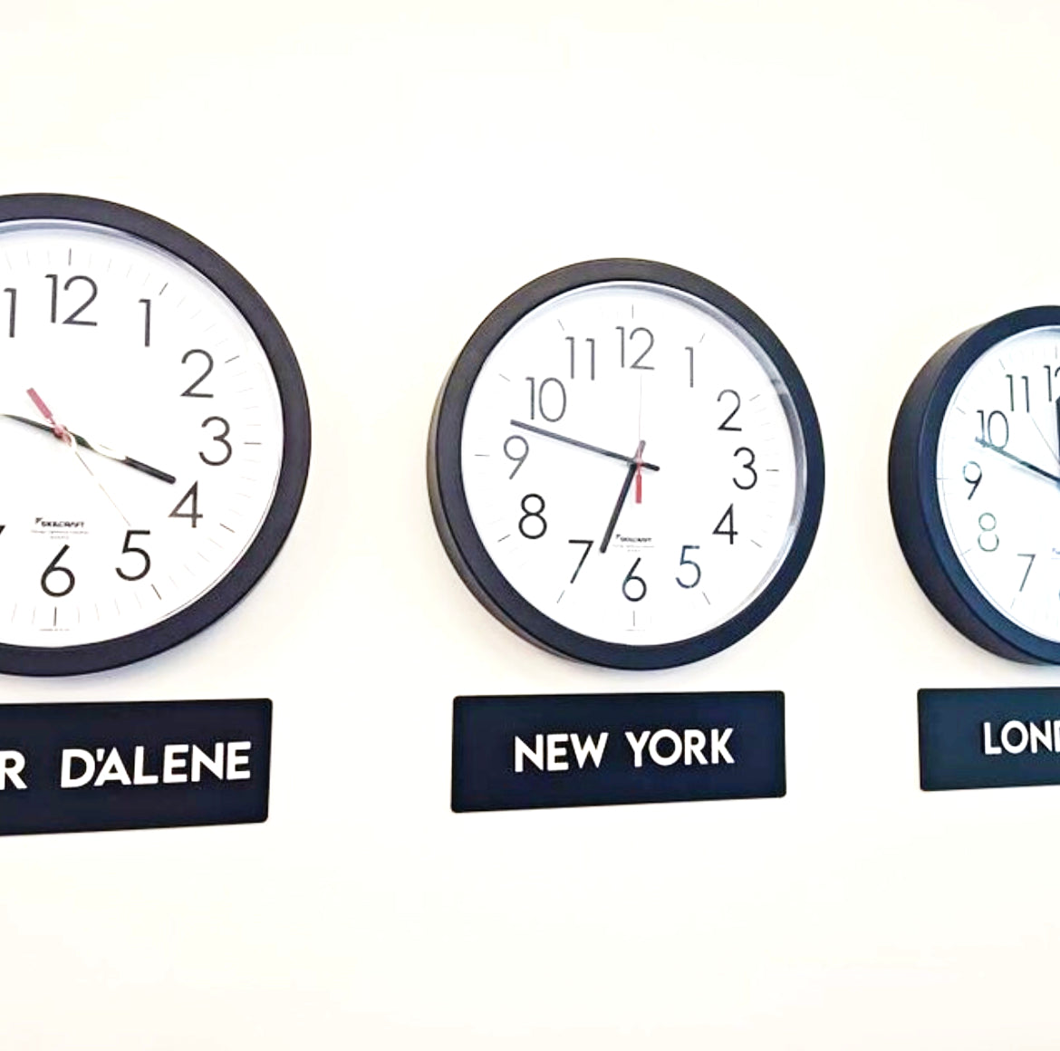 Three wall clocks labeled for different time zones on a white wall.
